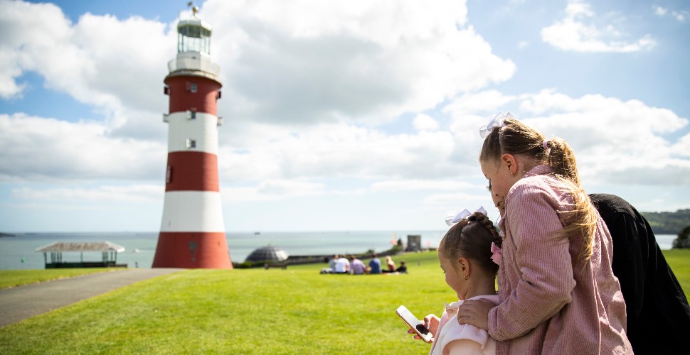 A family stood on Plymouth Hoe, with Smeaton's Tower in the background, look at the Plymouth Trails app on their phone